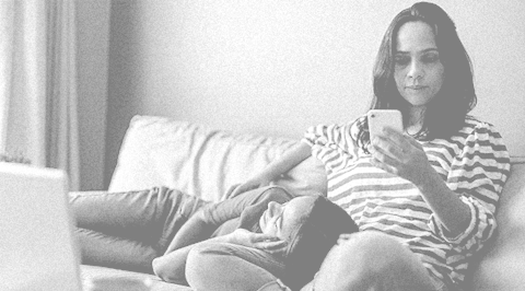 Mother sitting on the couch browsing on her phone while her teenage daughter is resting in her lap