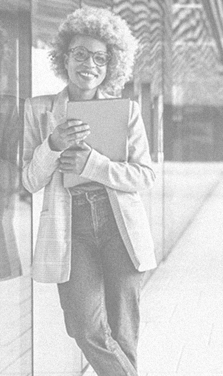 Colored woman wearing a long mens style blazer looking happy and confident leaning against glass building and holding a book.